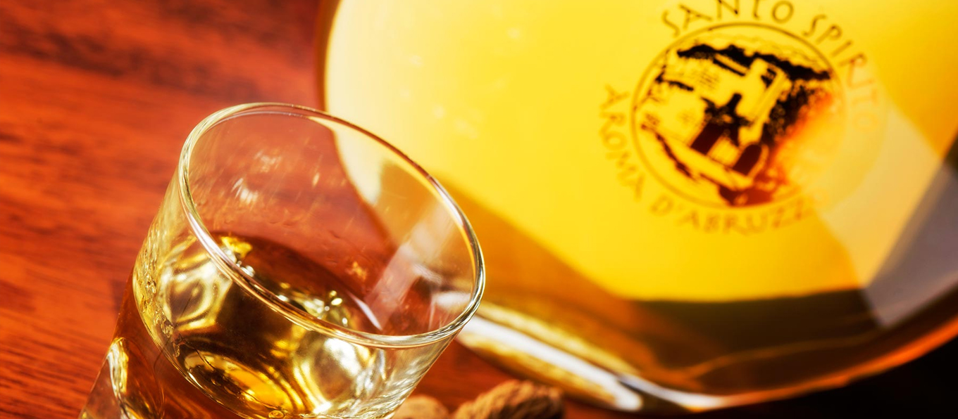 Bicchiere contenente liquore ambrato, poggiato su tavolo di legno e vicino una bottiglia trasparente con marchio Santo Spirito